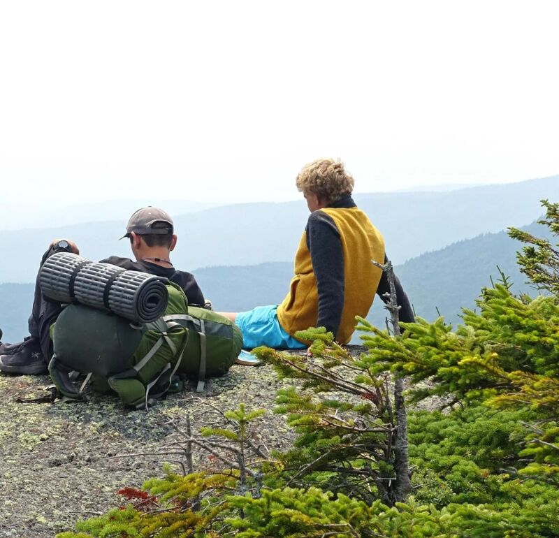 Two hikers are sitting on a rocky outcrop, taking in the scenic mountain view. One hiker, wearing a yellow vest, faces away from the camera, while the other, with a backpack and cap, is partially visible. The landscape features layers of distant mountains fading into the horizon, with some greenery in the foreground. The sky is overcast, creating a soft, diffused light over the scene.